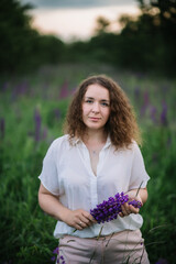 Young woman stands in white shirt in field of purple and pink lupins. Beautiful young woman with curly hair outdoors on a meadow, lupins blossom. Sunset or sunrise, bright evening light