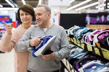 Mature man and woman standing in salesroom of appliance shop and choosing clothes iron. Woman pointing finger.