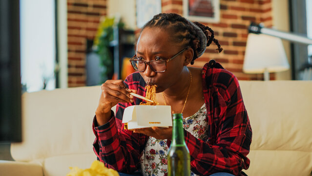 Female person using chopsticks to eat asian food, laughing at favorite film on television. Young woman eating noodles in delivery box and drinking beer, having fun at television. Handheld shot.