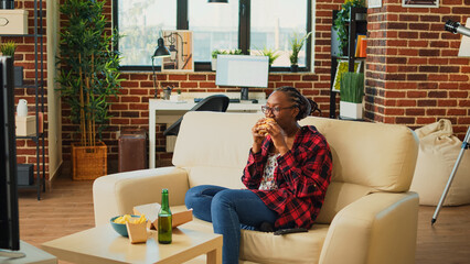 Young woman enjoying cheeseburger and beer at home, ordering fast food takeaway meal and watching...