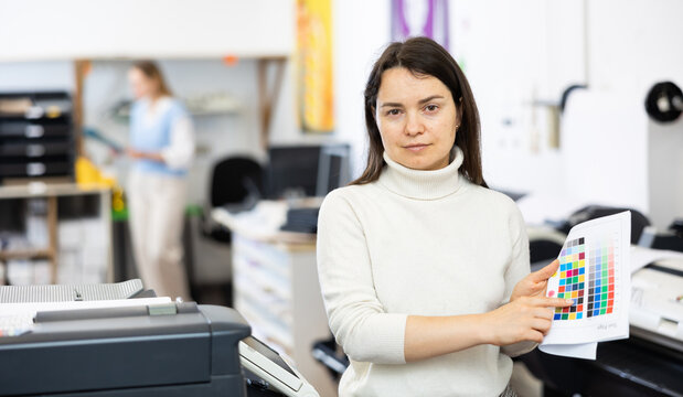 Portrait of positive woman printing office worker holding colour test page for printer and pointing finger.