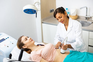 Confident female beautician performing body ultrasound cavitation procedure for european woman at aesthetic clinic
