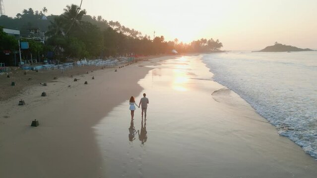 Couple holding hands walks on tropical beach along tide by ocean at sunrise front view drone footage. Boyfriend and girlfriend with coconuts walk at seaside on sand near water talk at dawn aerial view