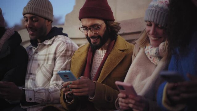 Group of cheerful young smile diverse people using cell phones. Happy friends addicted to social media typing mobile at night sitting outdoors in winter. Online relationships in millennial generation.