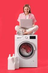Pretty young woman with modern laptop sitting on washing machine against red background
