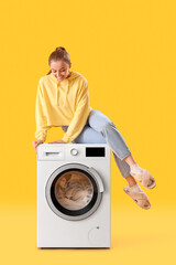 Pretty young woman sitting on washing machine against yellow background