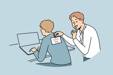 Man sticks kick me sticker on back of colleague working with laptop to make prank and laugh. Cheerful student guy jokes with friend and asks others not to talk about uplifting prank