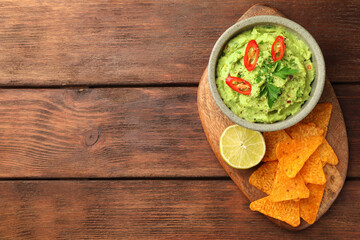 Bowl of delicious guacamole, lime and nachos chips on wooden table, top view. Space for text