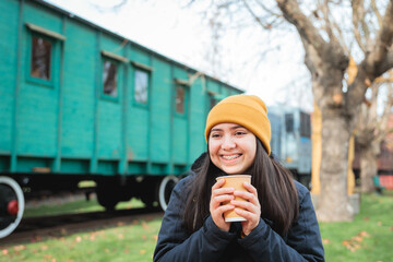 Cosy Vibes: Latina Girl, Dressed in Winter Clothing, Indulging in a Takeout Coffee at the Railway Park