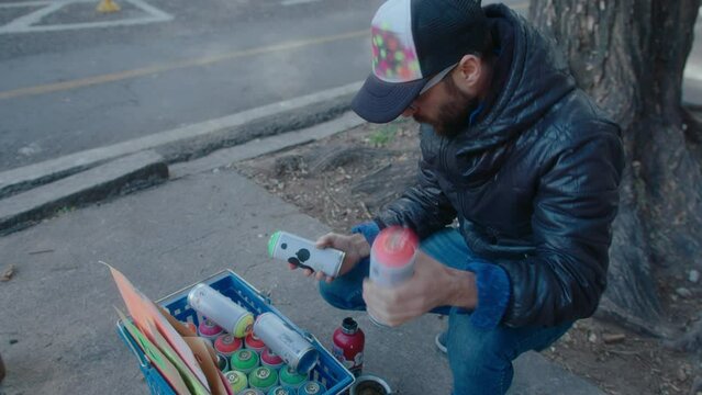 Graffiti artist taking spray paint from box with painting supplies, tossing bottle, preparing to draw outdoors on city street