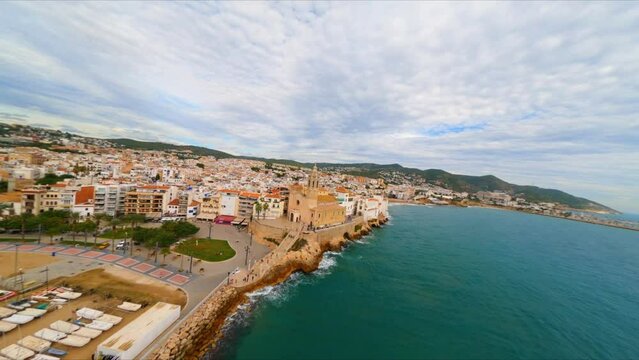 FPV aerial drone flying along the coast of Sitges, Spain with a view of Sant Bartomeu i Santa Tecla church.