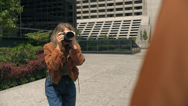 Young blonde woman taking photography of model on the city background.Photographer lady using camera for photoshoot.Attractive lady holding mirrorless photo camera looking on screen.