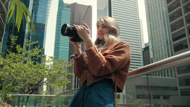 Young blonde woman taking photography modern city buildings.Photographer lady using camera for photoshoot.Photoshoot education.Attractive lady holding mirrorless photo camera looking on screen.