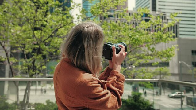 Young blonde woman taking photography modern city buildings.Blonde lady using camera for photoshoot.Attractive lady holding mirrorless photo camera looking on screen.