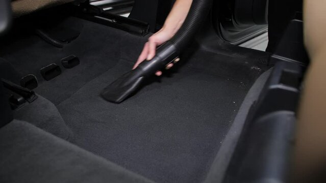 Removal of garbage on carpets in the car interior using a vacuum cleaner. A woman's hand controls the hose. Black interior and light seats.