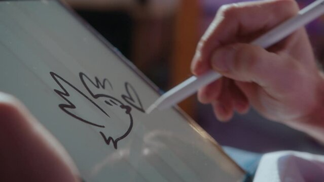 Hands of talented illustrator drawing cartoon style bird on a touchscreen of a digital tablet with stylus pen. Close-up impersonal shot