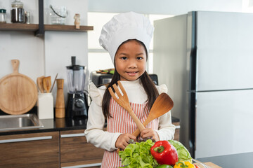 Asian Little chef girl cooking breakfast salad in the kitchen. Funny kids are preparing food with ingredient. Cute little asian girl a in chef with chef uniform