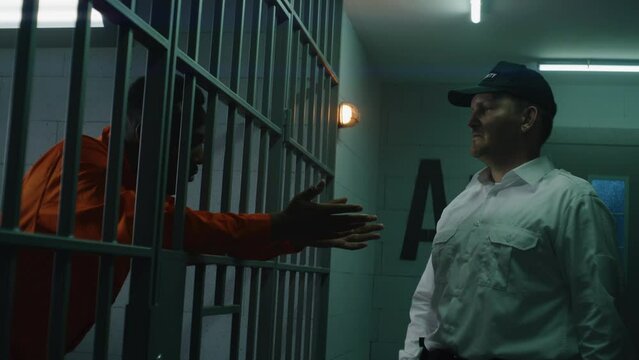 Warden with baton comes to prison cell, bullies prisoner and screams on him. Sad African American man serves imprisonment term in detention center. Murderer in orange uniform at correctional facility.