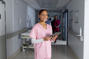 Fototapeta na wymiar Portrait of happy biracial female doctor wearing scrubs holding tablet in corridor at hospital