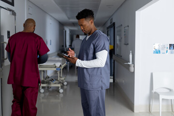 African american male doctor wearing scrubs using tablet in corridor at hospital