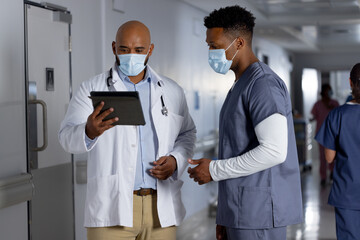 Diverse male doctors wearing face masks using tablet and talking in corridor at hospital