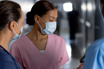 Diverse female doctors wearing face masks using tablet and talking in corridor at hospital