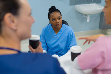 Diverse female doctors wearing scrubs drinking coffee, talking at reception desk at hospital