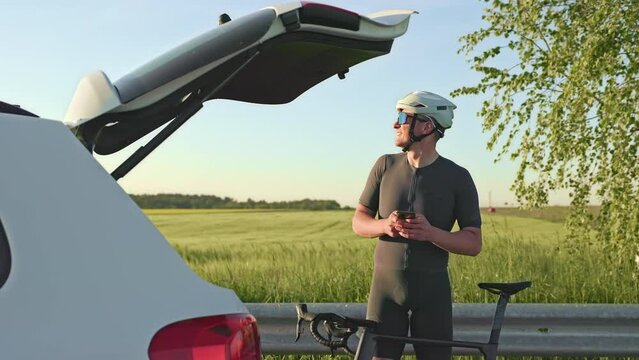 Young fit man with sports gearing utilizing smartphone while waiting with racing cycle near open car boot on sunny day. Delighted athlete checking email after finishing outdoor bike riding workout.
