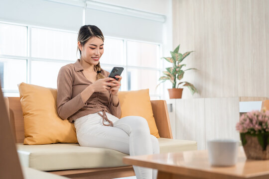 Beautiful young women using mobile phone sitting on sofa in living room. Using smartphone. Woman Shopping online store. Relax time at home