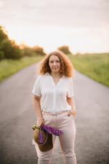 Young woman stands in white shirt on road with purple and pink lupins. Beautiful young woman with curly hair with bouquet of lupins. Sunset or sunrise, bright evening light