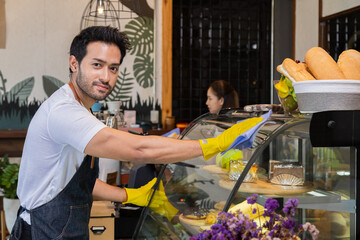 Small business owner multitask lifestyle concept, Asian coffee shop owner man or worker cleaning bakery cabinet for opening, multitask of entrepreneur wearing apron looking camera while working
