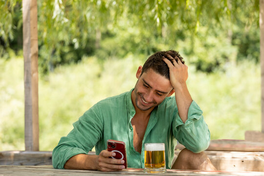 Young, handsome guy in the middle of summer sitting at a table with a beer in nature while looking at his phone smiling. .