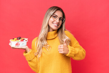 Young pretty Uruguayan woman holding a bowl of fruit isolated on red background giving a thumbs up gesture