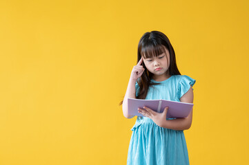 Fototapeta premium An adorable young Asian girl is reading a book, standing against an isolated yellow background