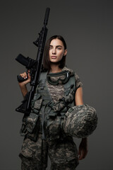 Stunning female soldier wearing a military uniform holding an automatic rifle against a grey...