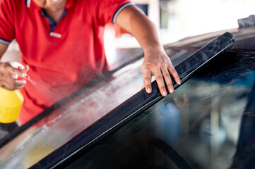 Male auto specialist worker hand spraying soapy solution while rolling car window film on front windscreen glass surface. Car side window film removal and tinting installation.  - 631010112