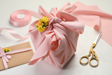 Furoshiki technique. Gift packed in pink fabric, card, flowers and scissors on white table, closeup