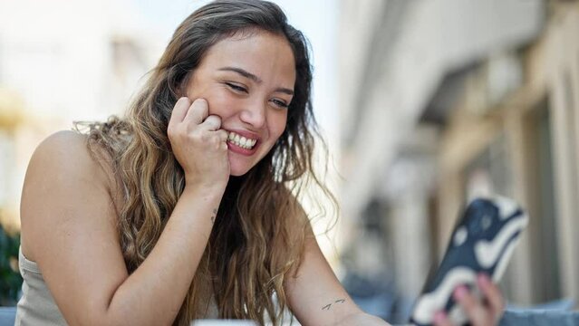 Young beautiful hispanic woman using smartphone sitting on table smiling at coffee shop terrace