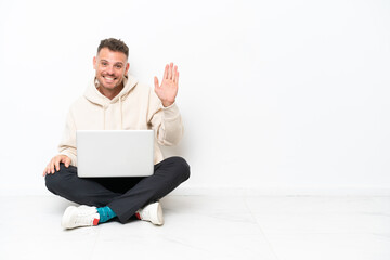 Young caucasian man with a laptop sitting on the floor isolated on white background saluting with hand with happy expression