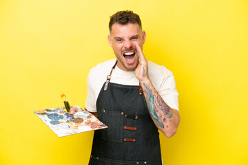 Young artist caucasian man holding a palette isolated on yellow background shouting with mouth wide...