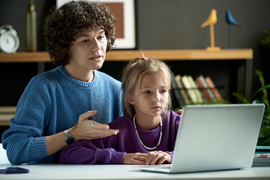 Mother explaining school material to her daughter pointing at laptop while they sitting at desk during their study
