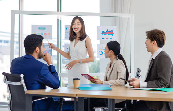 Millennial Asian professional successful businesswoman lecturer presenter in formal wears standing using pencil pointing presenting infographic paperworks documents on glass board in meeting room