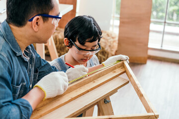 Happy Asian father and son carpentry wearing safety glasses and gloves. Father teaches his son to measure wood by measuring tape and marking with a pencil. Home workshop and Carpentry family concept.