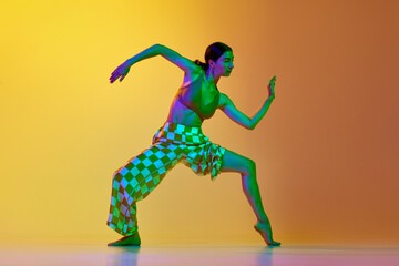 Artistic, dynamic image of young woman in motion, training, dancing against gradient yellow orange background in neon light. Concept of modern dance style, hobby, art, performance, lifestyle, ad