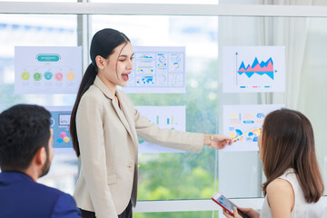Obraz premium Millennial Asian professional successful businesswoman lecturer presenter in formal wears standing using pencil pointing presenting infographic paperworks documents on glass board in meeting room