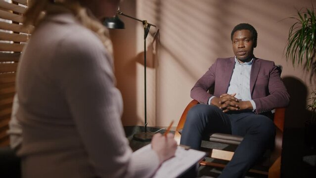 Female journalist with blonde hair interviewing successful businessman in room with closed blinds. Handsome man in elegant suit sitting in chair and talking about development of his business.