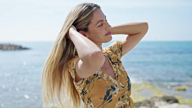 Young blonde woman smiling confident combing hair at seaside