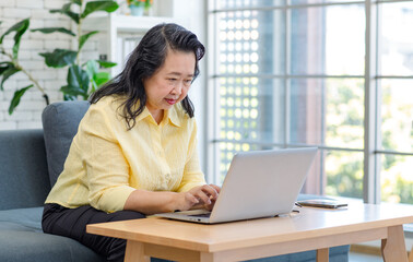 Fototapeta premium Asian old senior pensioner chubby fat housewife sitting on cozy sofa while surfing browsing internet online via laptop computer in living room.