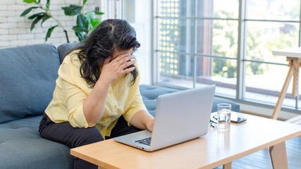 Asian old senior pensioner chubby fat housewife sitting on cozy sofa headache attack while surfing browsing internet online via laptop computer.