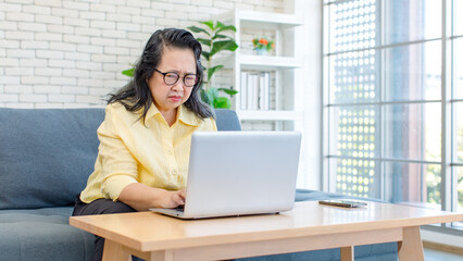 Asian old senior pensioner chubby fat housewife sitting on cozy sofa  while surfing browsing internet online via laptop computer in living room.